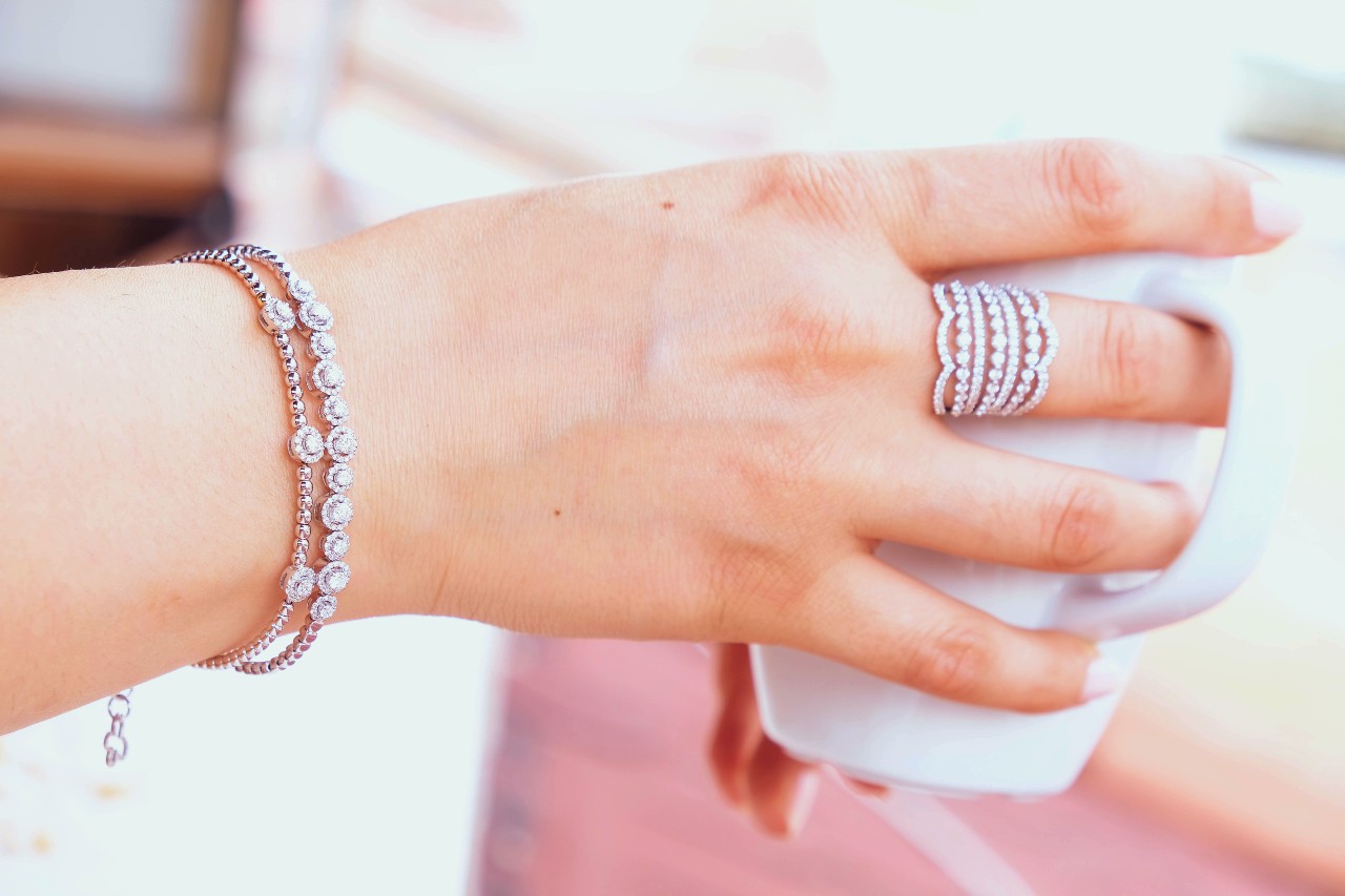 A hand adorned with a diamond bracelet and a multi-row diamond ring holds a white cup.