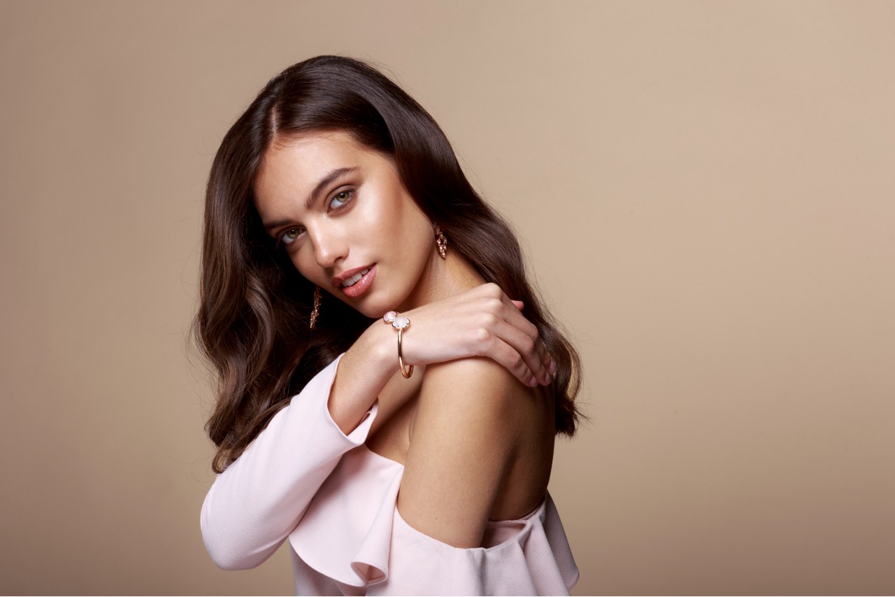 A woman with wavy hair poses in a blush off-shoulder outfit, showcasing a rose gold floral bracelet and matching earrings.