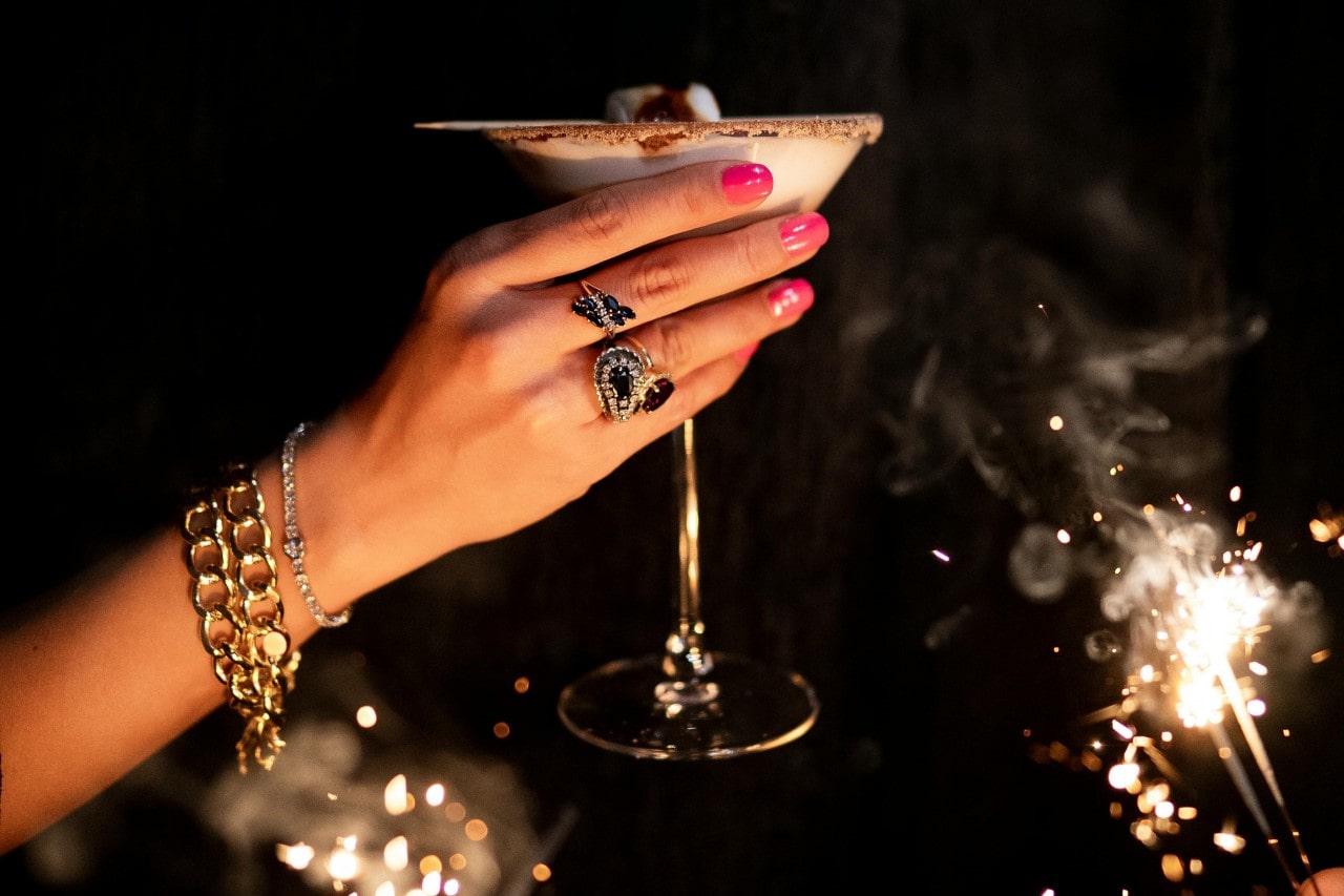 A woman toasts with her smores martini while wearing gold chain bracelets, a diamond tennis bracelets, and a couple statement sapphire and diamond rings with sparklers burning in the background.