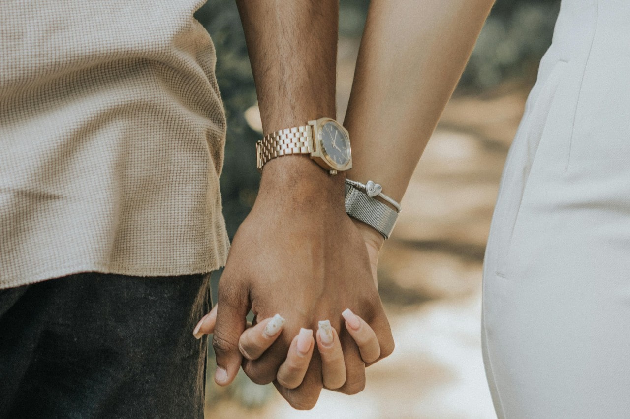 A close up of a couple holding hands with a man wearing a yellow gold metallic wristwatch and a woman wearing a silver bracelet with a heart charm.