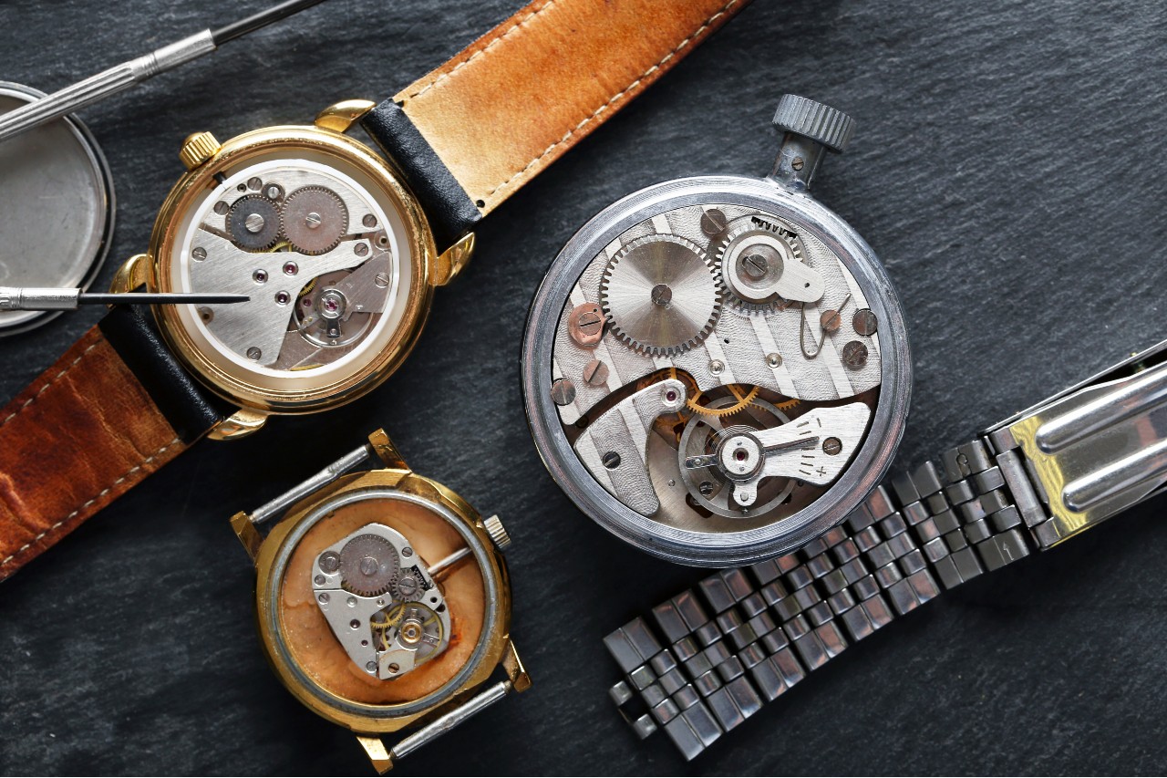 Close-up view of mechanical watch movements being repaired with precision tools on a dark work surface.