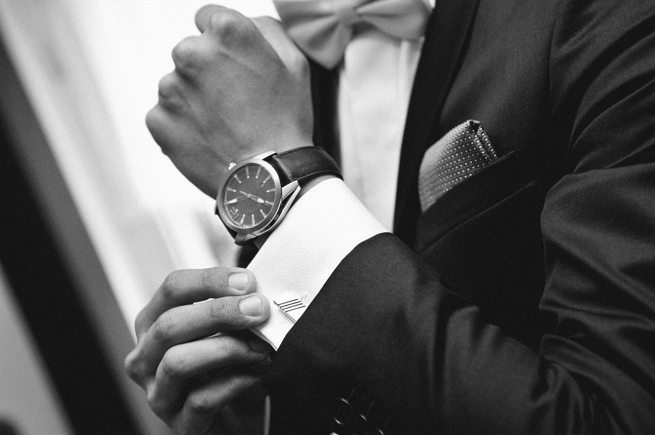 A black and white photo of a man in a suit fixing his cuffs, showcasing black leather wristwatch