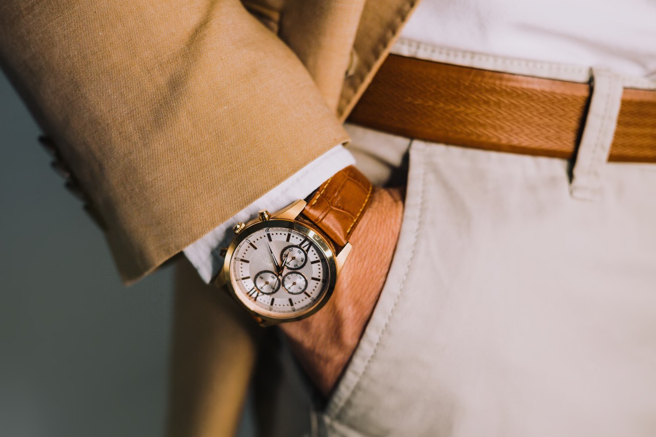 A close up of a man’s hand in a pocket of his beige trousers, showcasing a wristwatch with leather bracelet.