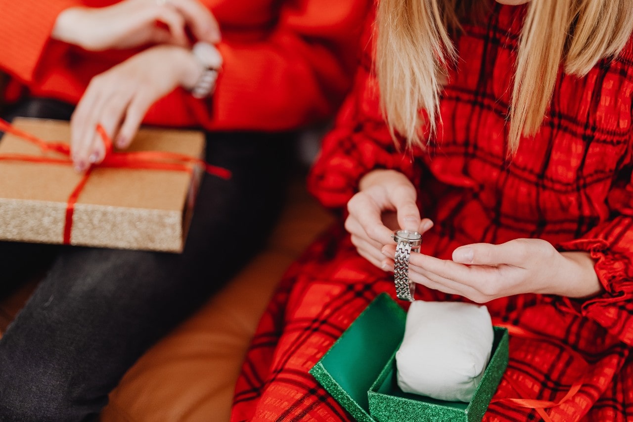 A woman in a red plaid dress admires a silver wristwatch she received from a green gift box while another person holds a wrapped present.