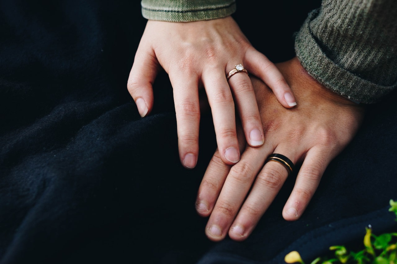A close up of a couple’s hands on a blue blanket, showcasing rose gold diamond wedding engagement ring and dual tone men’s wedding band.