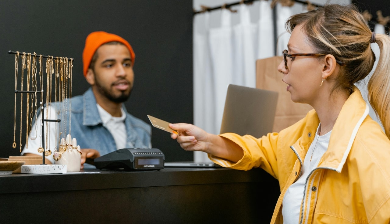 A woman in a yellow jacket pays with a credit card at a jewellery store counter while a cashier assists her.