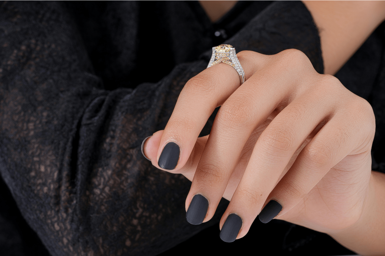 A close up of a halo engagement ring encrusted with white and yellow diamonds on a woman’s hand with black nails and in front of her black blouse.