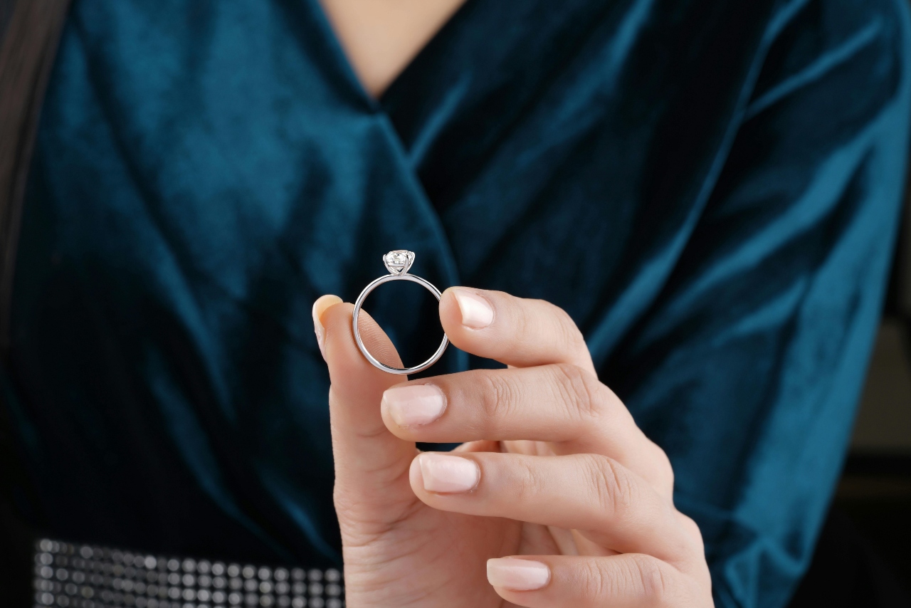 A close-up of a person in a dark teal outfit holding a solitaire diamond engagement ring between their fingers.