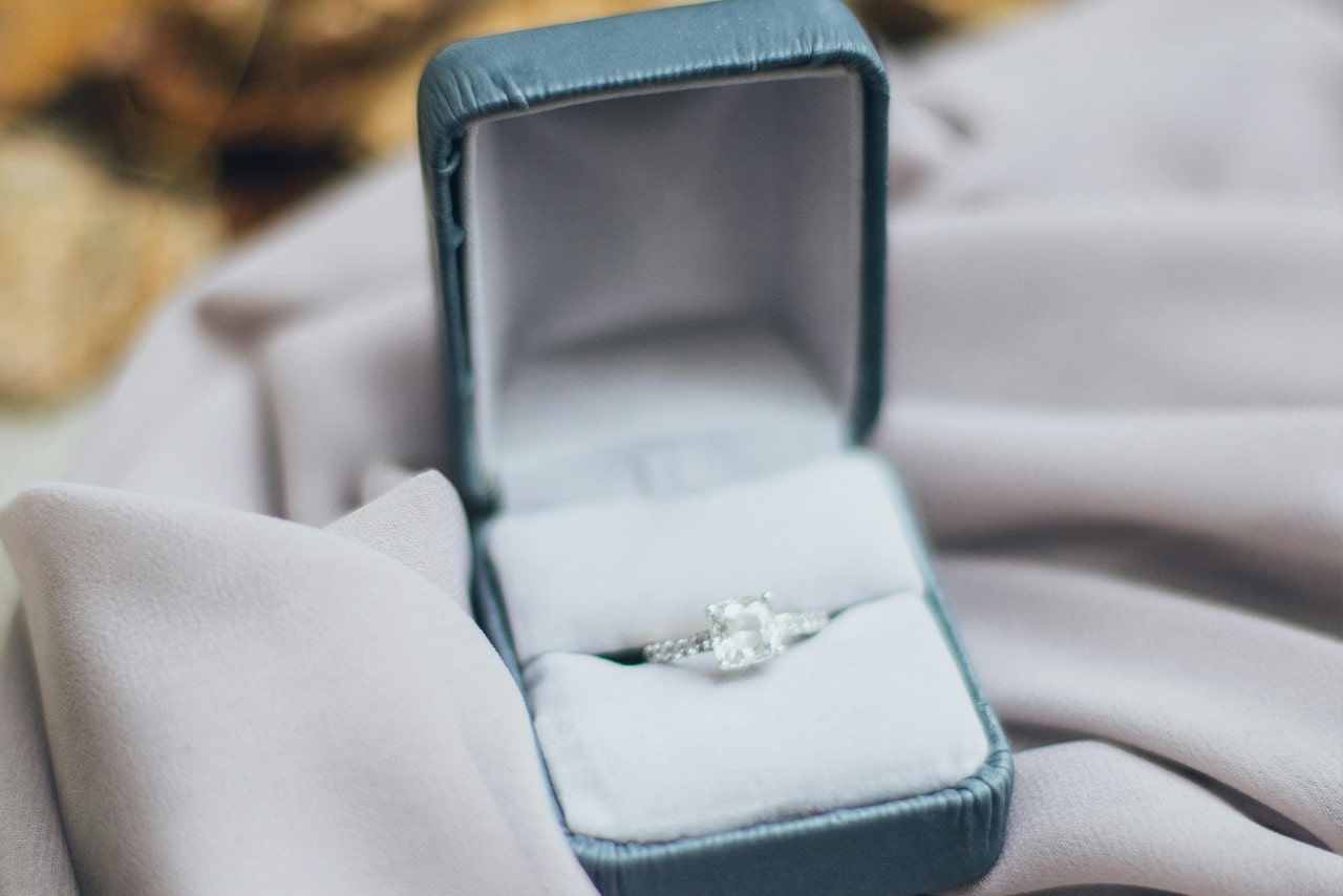 A close up of a white gold princess cut engagement ring in a green velvet box on a piece of white fabric.