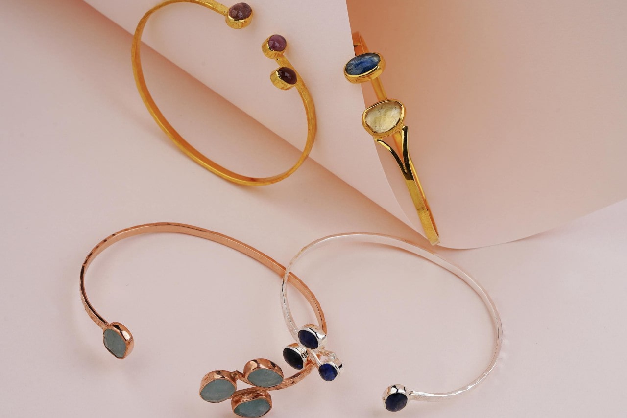 A collection of colourful gemstone bangles in gold, silver, and rose gold displayed on a soft pink background. A collection of colourful gemstone bangles in gold, silver, and rose gold displayed on a soft pink background.