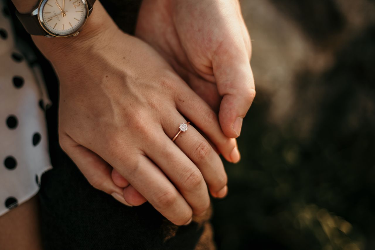 A close-up of a rose gold solitaire engagement ring on a woman’s hand held by a man’s hand against a blurry background of her polka dot skirt. A close-up of a rose gold solitaire engagement ring on a woman’s hand held by a man’s hand against a blurry background of her polka dot skirt.
