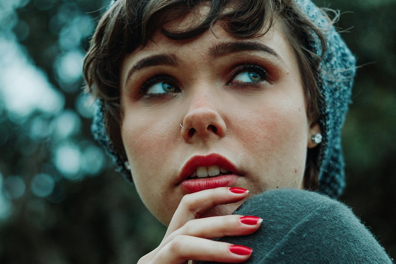 A young woman with short hair and a beanie thoughtfully gazes upward, wearing an elegant pair of diamond stud earrings.