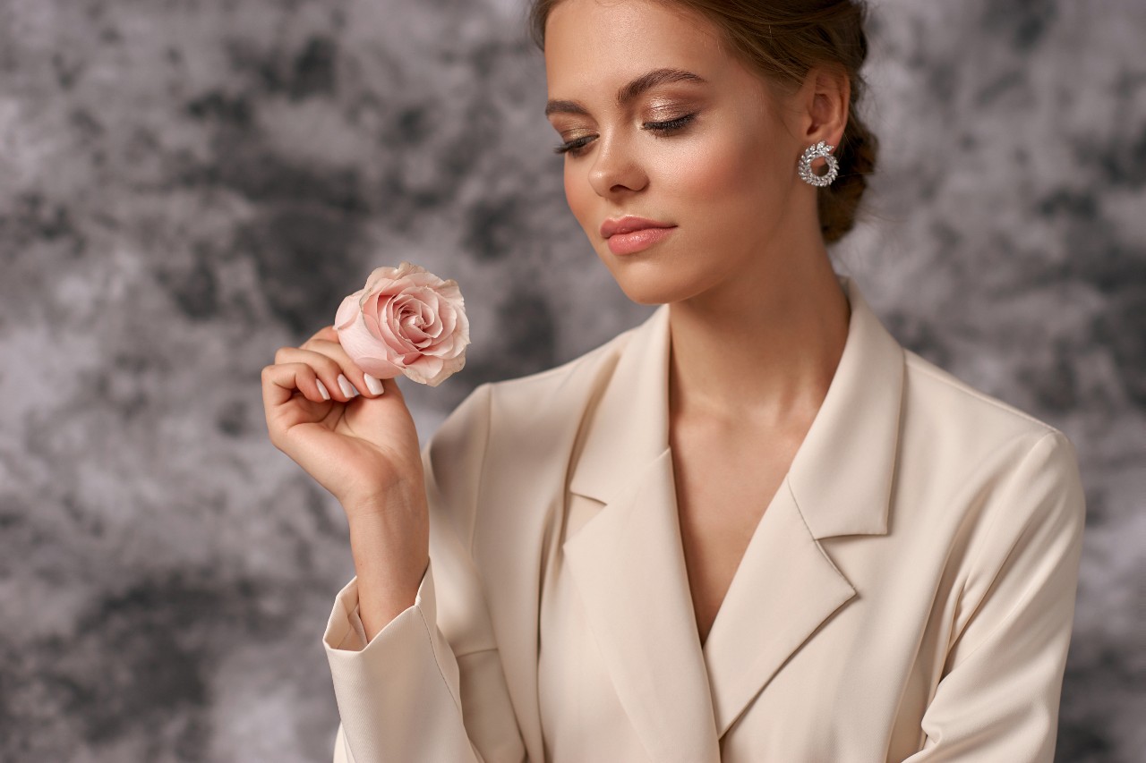 An elegant woman in a beige blazer and sparkling earrings holding a pink rose, eyes closed, against a soft gray background.