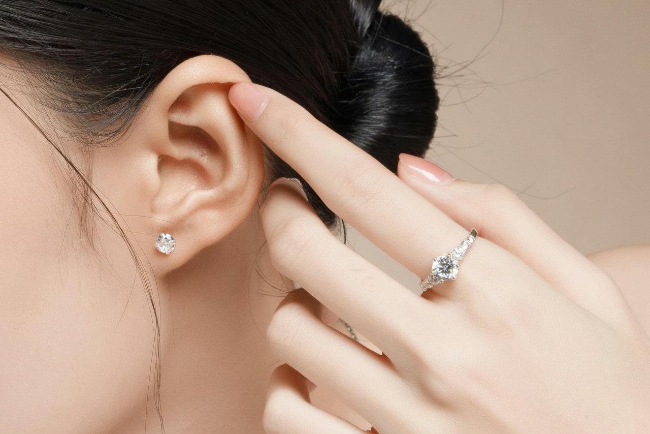 A close-up of white gold diamond studs on the woman’s ear and matching ring on her finger.
