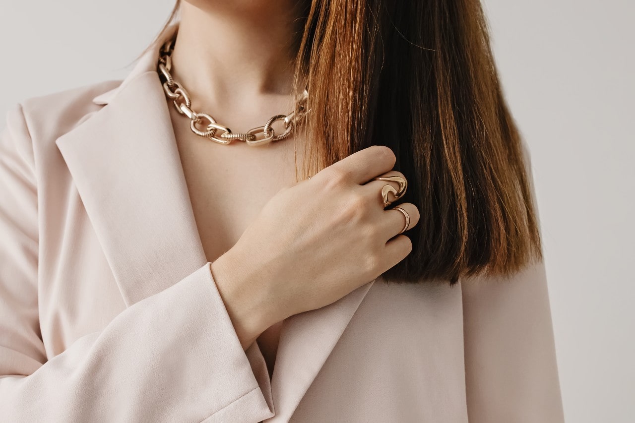 A close-up of a woman in beige blazer with a chunky yellow gold chain necklace and matching polished rings.