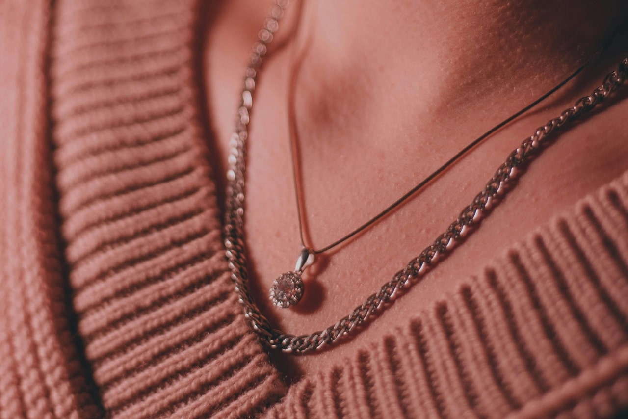 A close up of two silver necklaces: one is a halo gemstone pendant and the other is a chain necklace on a woman’s neck who is wearing a pink sweater. A close up of two silver necklaces: one is a halo gemstone pendant and the other is a chain necklace on a woman’s neck who is wearing a pink sweater.