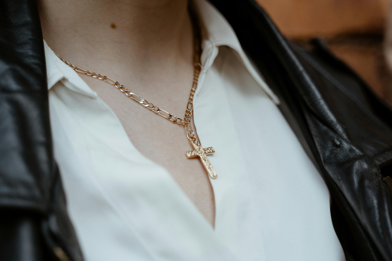 A close up of a yellow gold chain cross necklace on a person who wear white blouse and black leather jacket. A close up of a yellow gold chain cross necklace on a person who wear white blouse and black leather jacket.