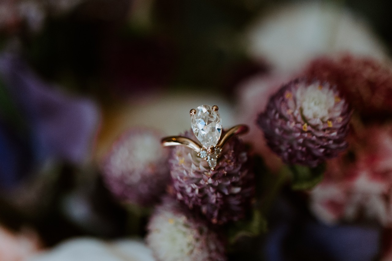 A pear-shaped diamond engagement ring with a gold band is delicately perched atop purple clover flowers in a softly blurred floral background.