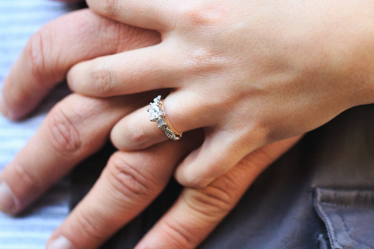 a pair of intertwined hands, one of which is wearing a mixed metal engagement ring