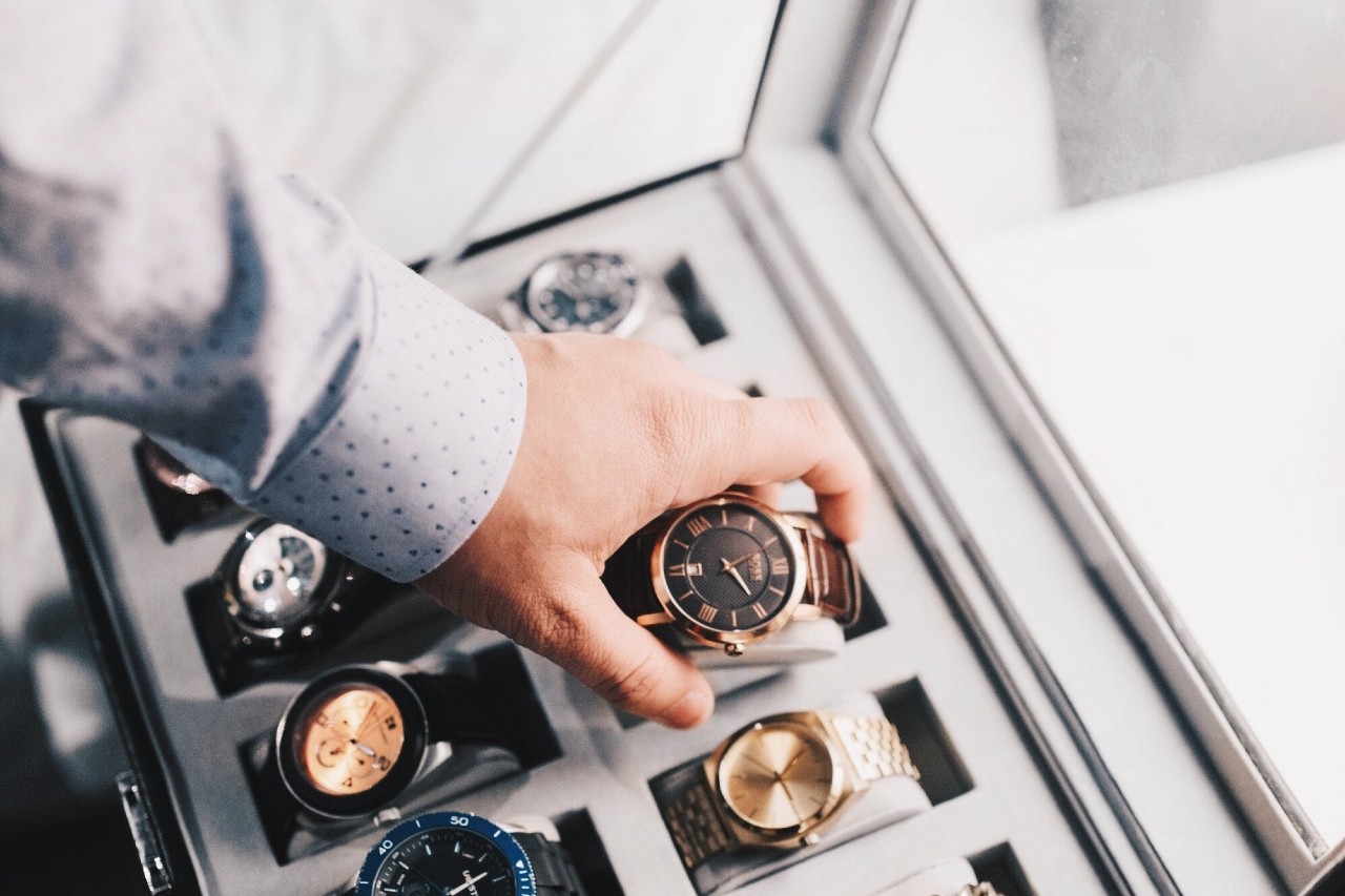 A person in a light blue shirt selects a yellow gold watch from a display case filled with various luxury timepieces. A person in a light blue shirt selects a yellow gold watch from a display case filled with various luxury timepieces.
