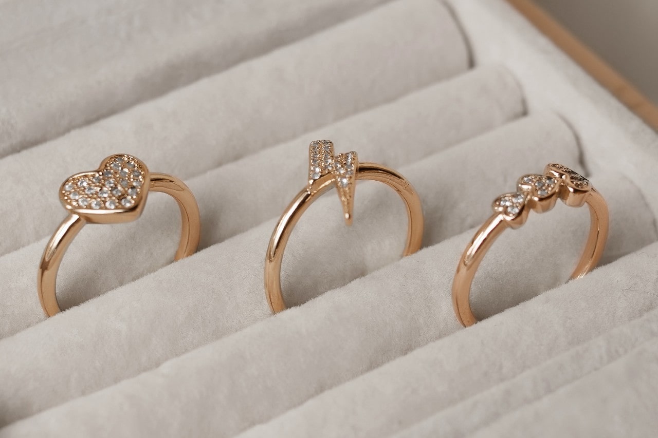 Three yellow gold rings with distinct designs — a heart, a lightning bolt, and triple teardrops— are arranged in a velvet-lined jewellery tray. Three yellow gold rings with distinct designs — a heart, a lightning bolt, and triple teardrops— are arranged in a velvet-lined jewellery tray.