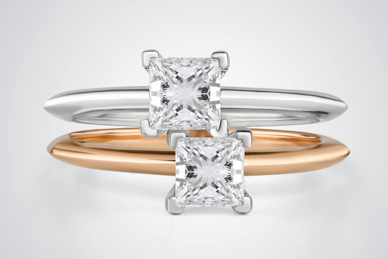 A close up of two solitaire princess cut engagement rings, one is white gold and the other is yellow gold. A close up of two solitaire princess cut engagement rings, one is white gold and the other is yellow gold.